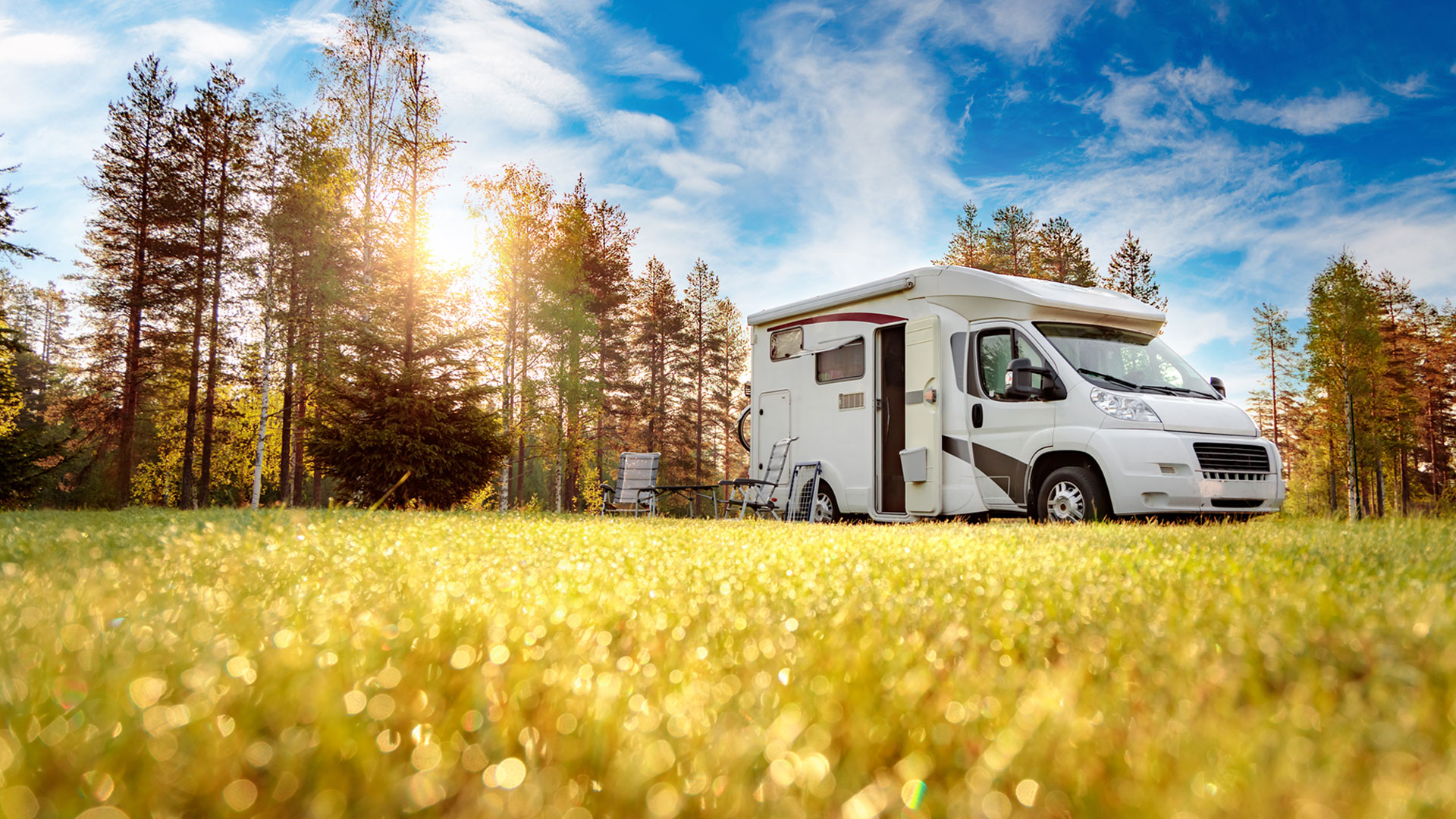 Weißes Wohnmobil auf sonniger Wiese am Waldrand mit Campingmöbeln unter blauem Himmel – Wohnmobil mieten bei Caravan24